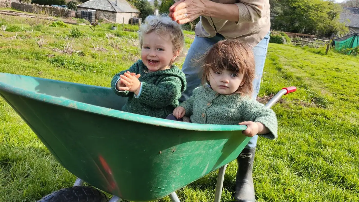 Two small children in green knitted cardigans sitting in a green wheelbarrow on the grass beside the plots.