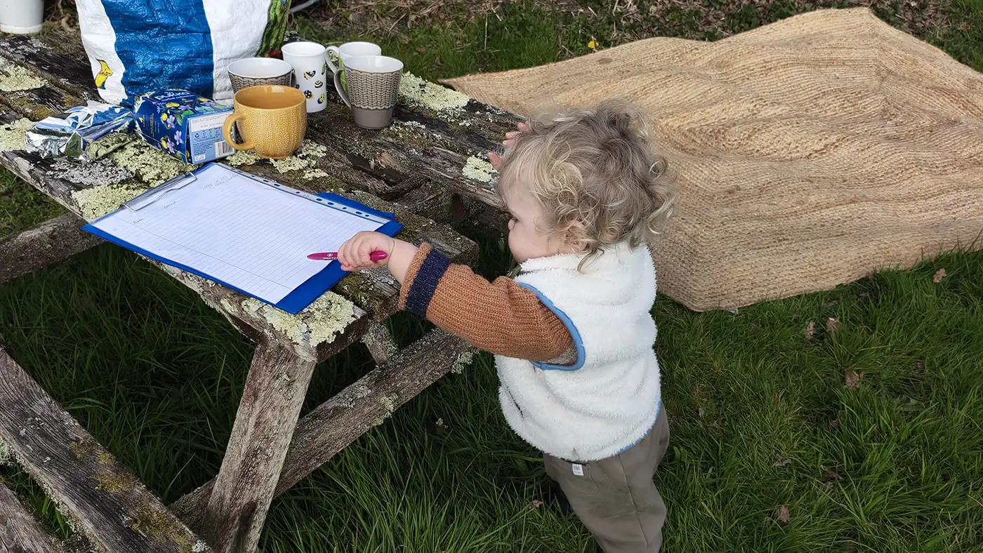 A toddler reaching up to a sign-up sheet on a clipboard, laid out on the lichen-covered picnic table with mugs of tea.